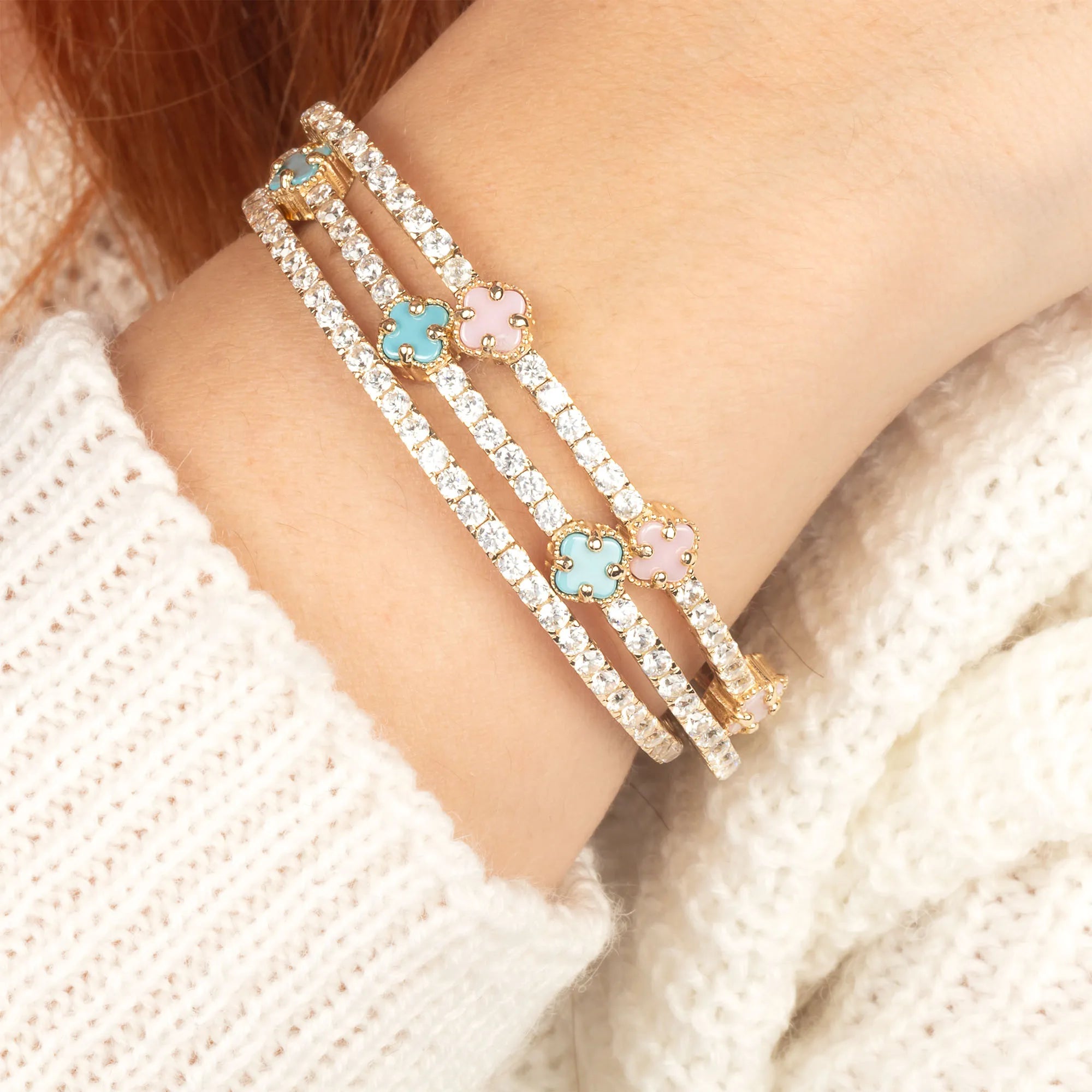 A wrist adorned with pastel blue and pink enamel clover diamond tennis bracelets in gold, showcasing a stylish and elegant look.
